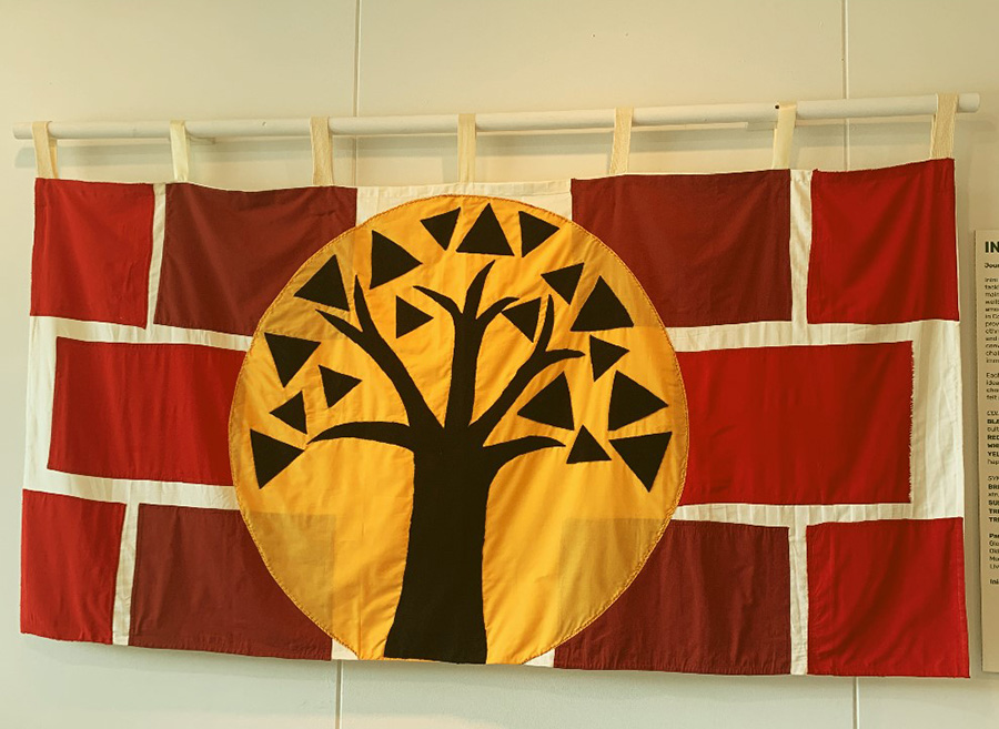 A flag with a white background and red squares on top. In the middle, there is a bright yellow circle, on top of it a black tree.