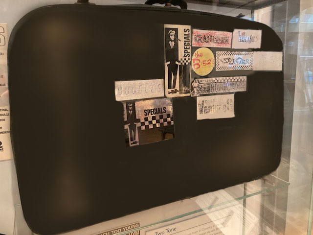A black vintage suitcase featuring various ‘The Specials’ stickers and other miscellaneous stickers, mostly black and white but also a faded red one and a yellow one.