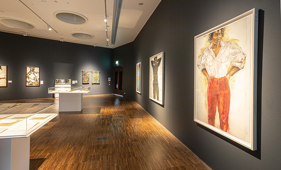 A photograph of an art exhibition. The walls are painted dark grey and the floors are wooden. Various works of art are hung on the walls and three white display cases with glass lids are visible in the space.