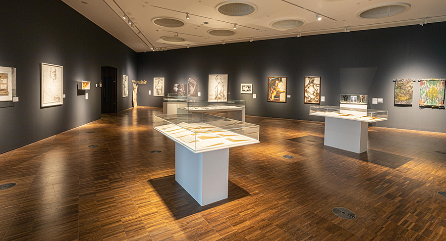 A wide-angle view of an art exhibition. The walls are painted dark grey and the floors are wooden. Various works of art are hung on the walls and four white display cases with glass lids are in the centre of the space.