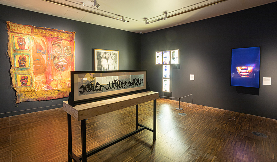 A photograph of an art exhibition. The walls are painted a dark grey and the floors are wooden. A textile work and a framed work are on one wall, with two screen-based works on the other wall. A wooden plinth with a mounted and framed transparent artwork 