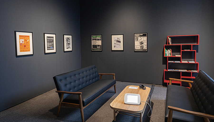 A photograph of a gallery space. The walls are painted dark grey and the floor is carpeted. There are six posters hung on the wall with a red bookshelf filled with books. In the space there are two sofas with a wooden table between them. There is a screen