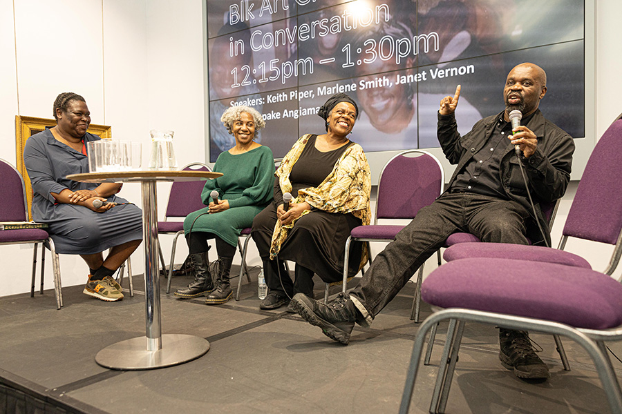 Four people sit on purple chairs on a raised stage in front of a screen. From left to right they are: Sepake Angiama, a Black woman wearing a navy dress; Marlene Smith, a Black woman wearing a green dress; Janet Vernon, a Black woman wearing a black dress