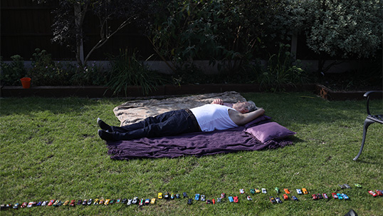 An old man in a white vest lying on a blanket, on the grass, in the garden. In the foreground, a long line of small child’s racing cars have been carefully arranged.
